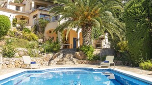 Vue sur une villa de style méditerranéen, avec un escalier en pierre menant à une entrée couverte, le tout entouré d’une végétation luxuriante et surplombé d’un ciel d’un bleu éclatant.