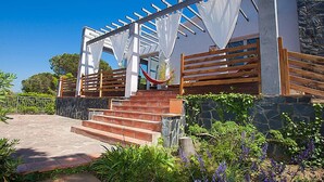 Un escalier en pierre mène à une terrasse en bois surmontée d’une pergola, le tout étant entouré d’une végétation luxuriante et de fleurs violettes.