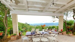 A covered patio with a table set for dining, surrounded by potted plants and white metal chairs, overlooking a scenic landscape. - Paradiso by Interhome (Perugia)