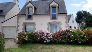 Una casa de dos pisos con fachada de piedra, dos ventanas visibles y un jardín con hortensias.