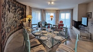 A dining area with a glass table set for six, surrounded by chairs. There is a sofa and a television in the background. - Les Rimains by Interhome (Cancale)
