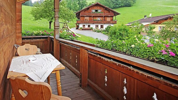 En træterrasse med et bord og stole, der ud til et landligt landskab med et stort hus og grøn vegetation.