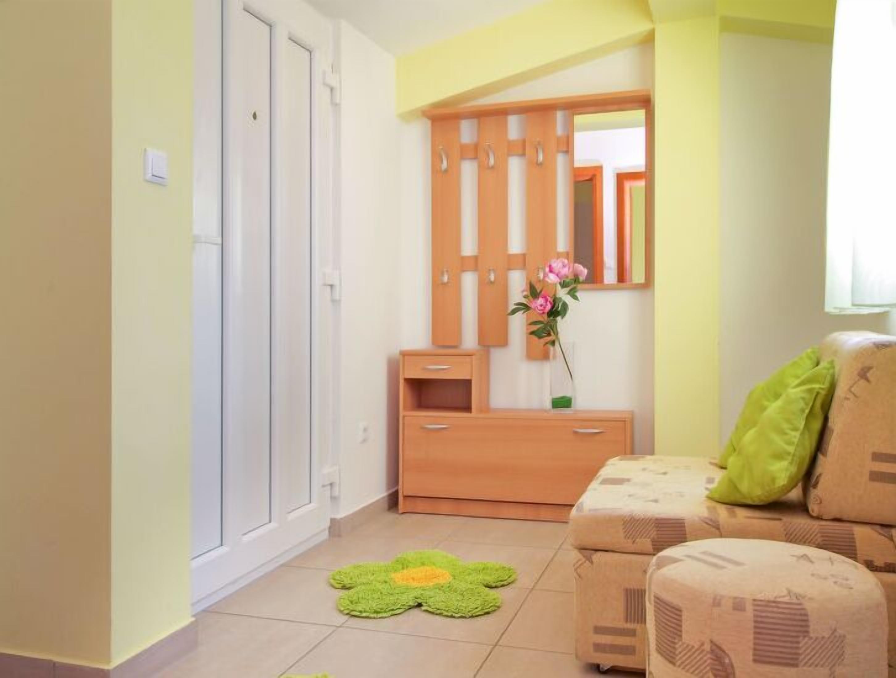 A room with a wooden wardrobe, a mirror, a small table with a vase of flowers, and a green cushion on a patterned sofa.