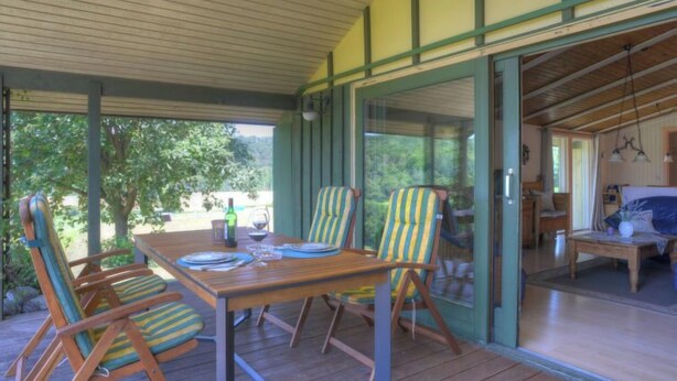 Un espace extérieur abrité, aménagé pour les repas, avec une table en bois et des chaises à rayures, de grandes fenêtres et une vue sur la verdure.