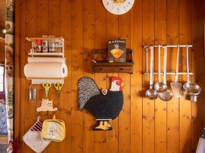 A kitchen with wooden walls, a clock, a shelf with kitchen utensils, a rooster decoration, and hanging kitchen tools.