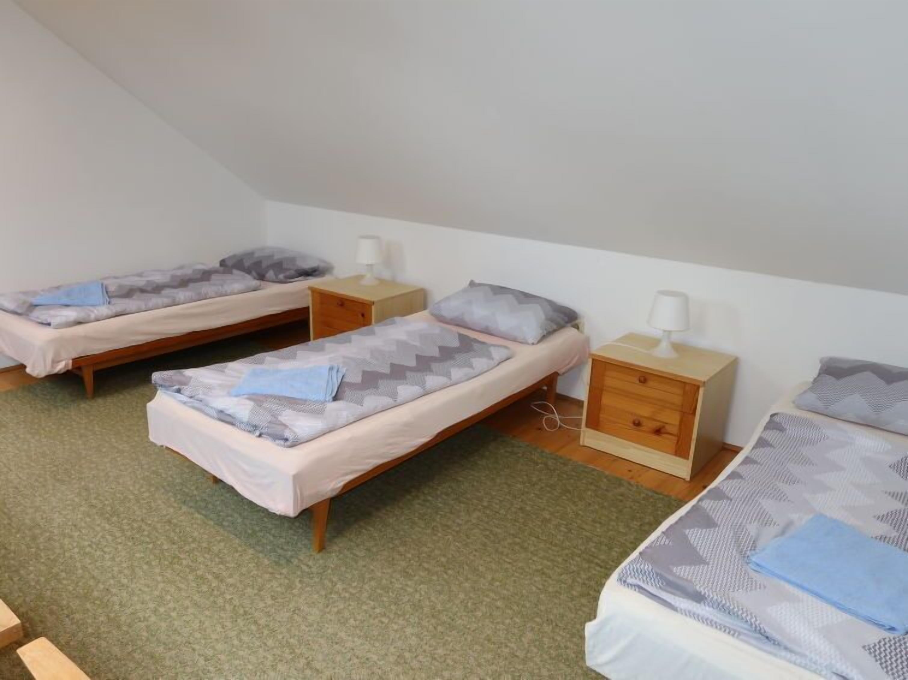 Una habitación tipo dormitorio con tres camas individuales, cada una con una mesita de noche y una lámpara. La habitación tiene techo inclinado y alfombra verde.