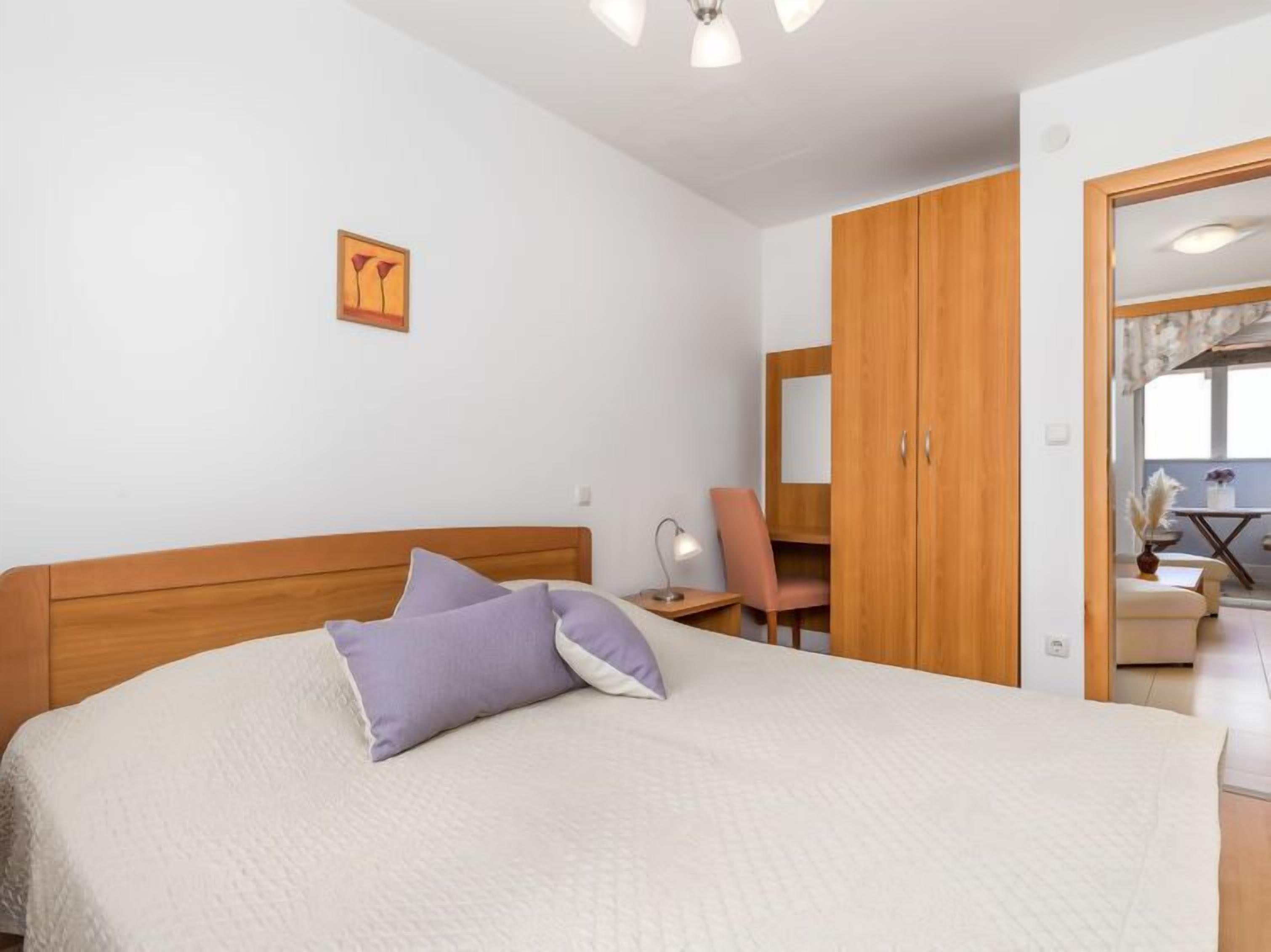 A bedroom with a large bed, a wooden wardrobe, a desk with a chair, and a window with curtains.
