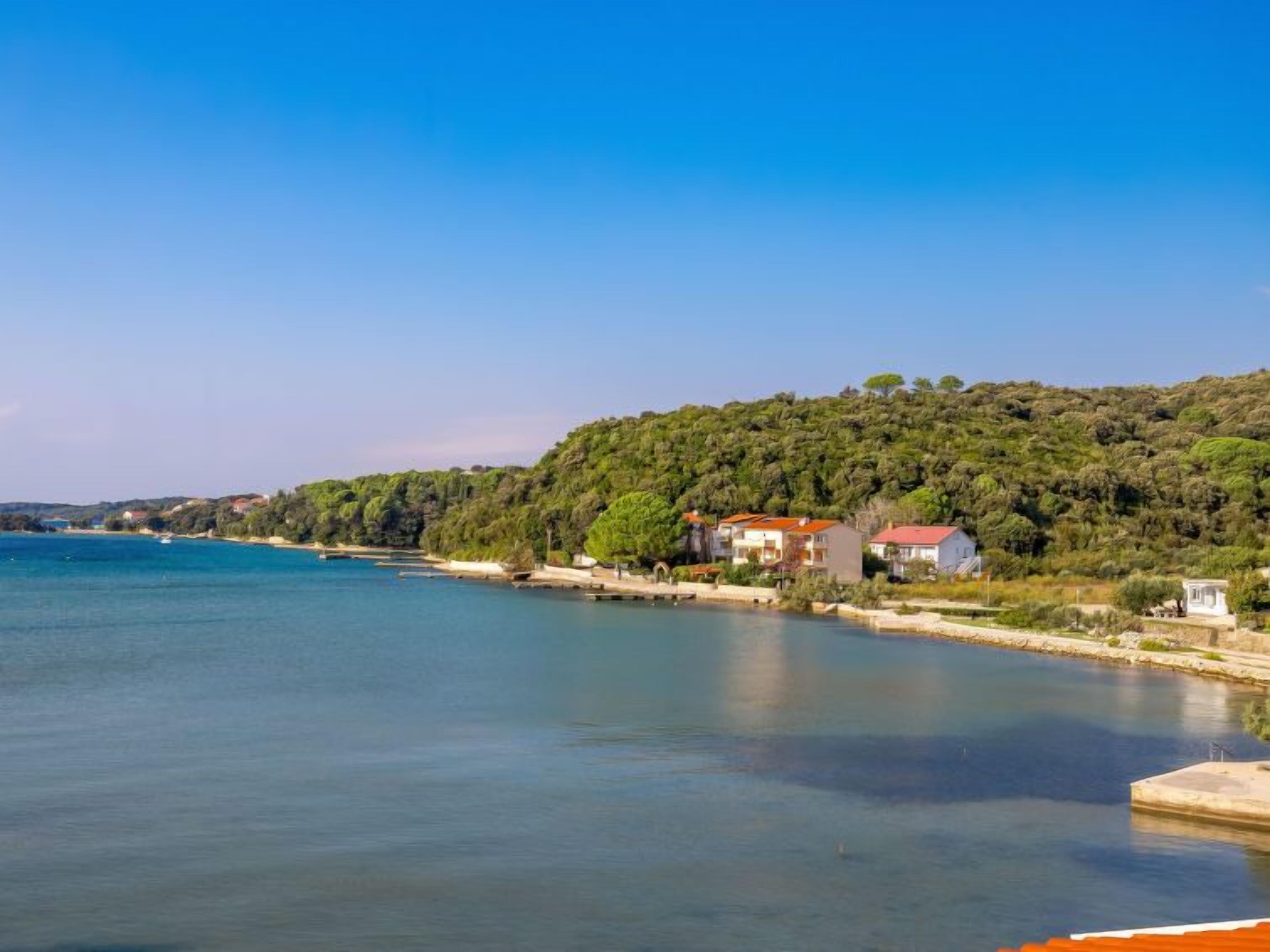 A coastal area with a clear view of the sea, a hillside with dense vegetation, and several buildings with red roofs.