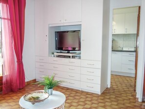 A room with a television on a white cabinet, a small table with a bowl and plant, and a kitchen area with white cabinets.