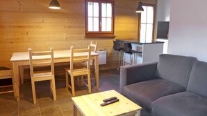 Una sala de estar acogedora con un sofá gris, una mesa de comedor de madera con sillas, una pequeña mesa de centro de madera y una zona de cocina con barra y sillas.