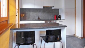 Una cocina moderna con encimera de granito, dos taburetes negros, armarios blancos y una lámpara colgante.