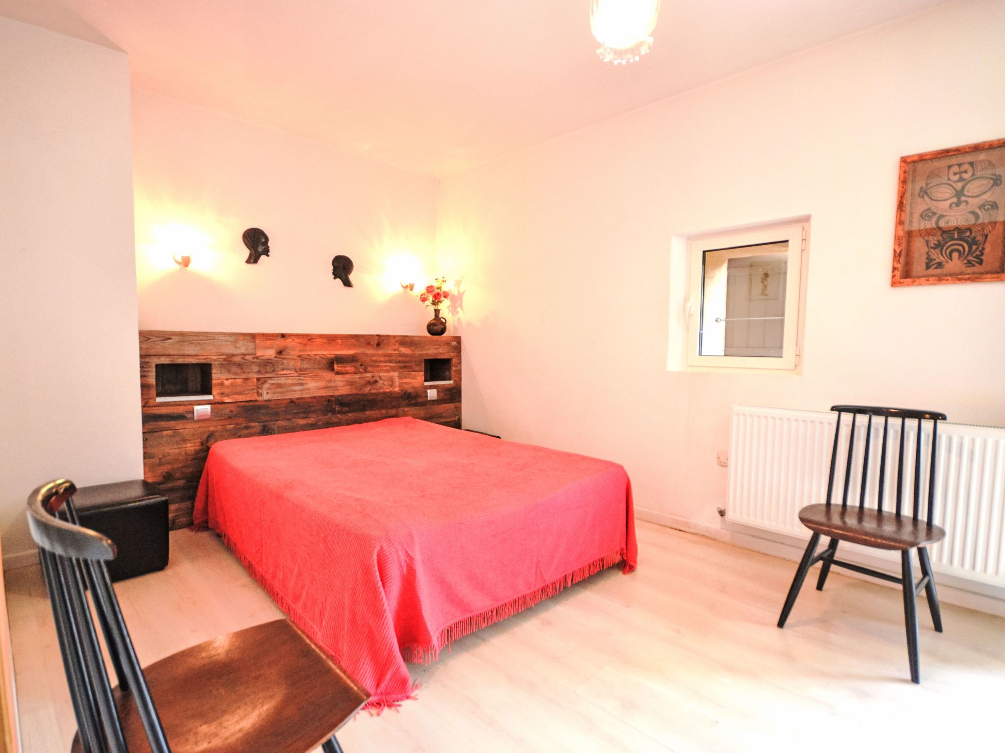 Een slaapkamer met een houten hoofdbord, een rode spreideken, een stoel, een nachtkastje en een raam met uitzicht.