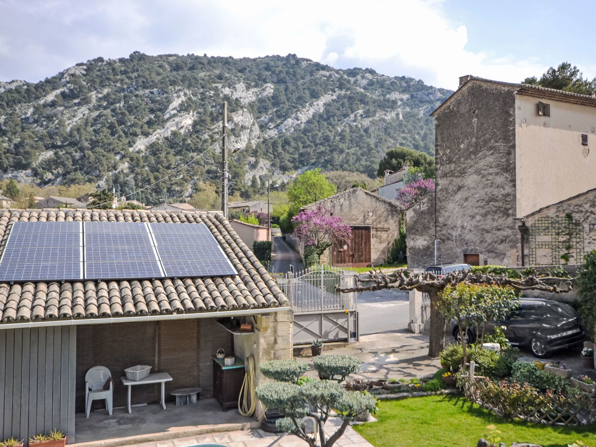 A rustic building with a tiled roof and solar panels, surrounded by a well-maintained garden and a mountainous backdrop.