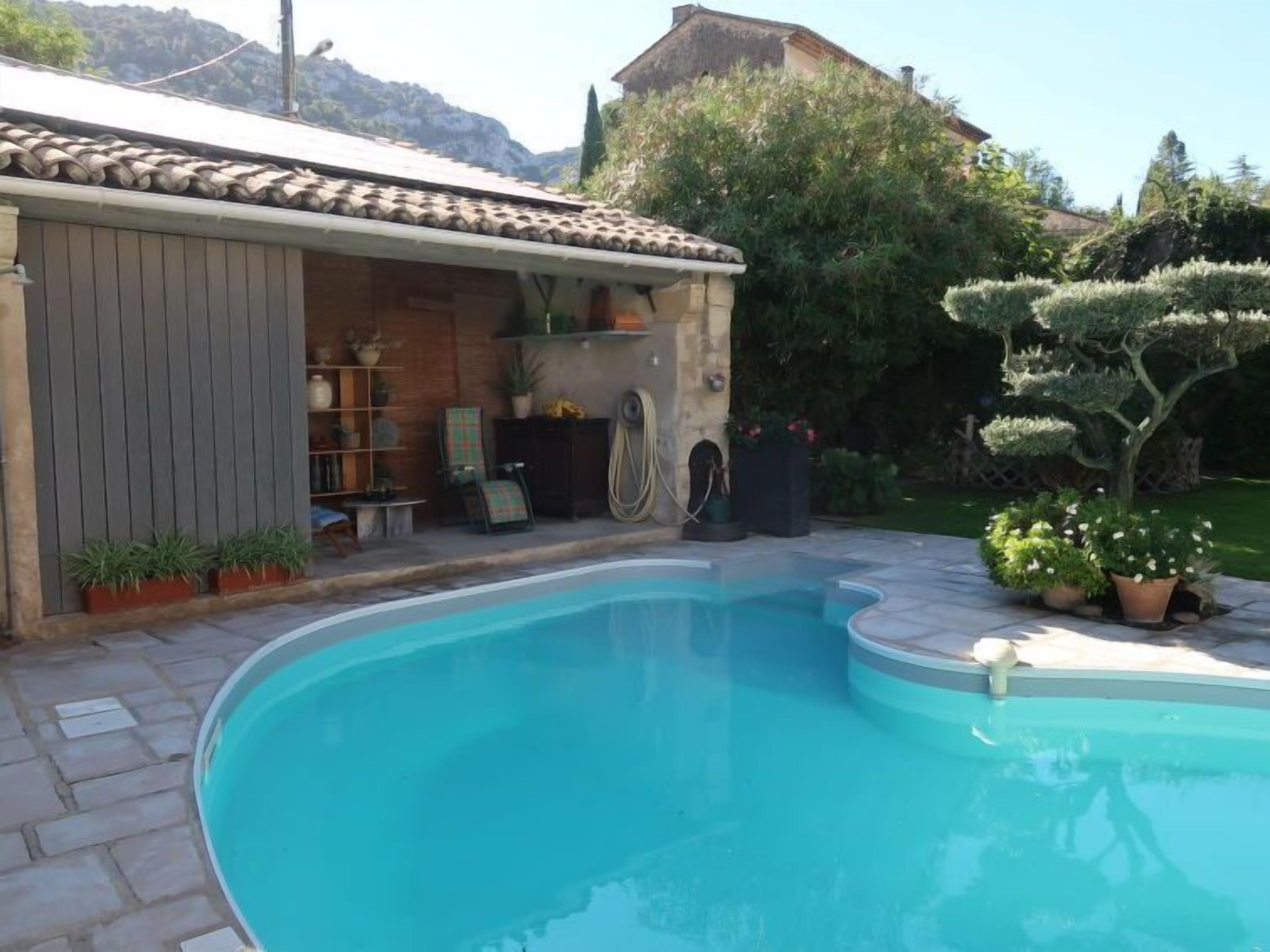 A swimming pool with a tiled deck, surrounded by potted plants and a small building with shelves and chairs.