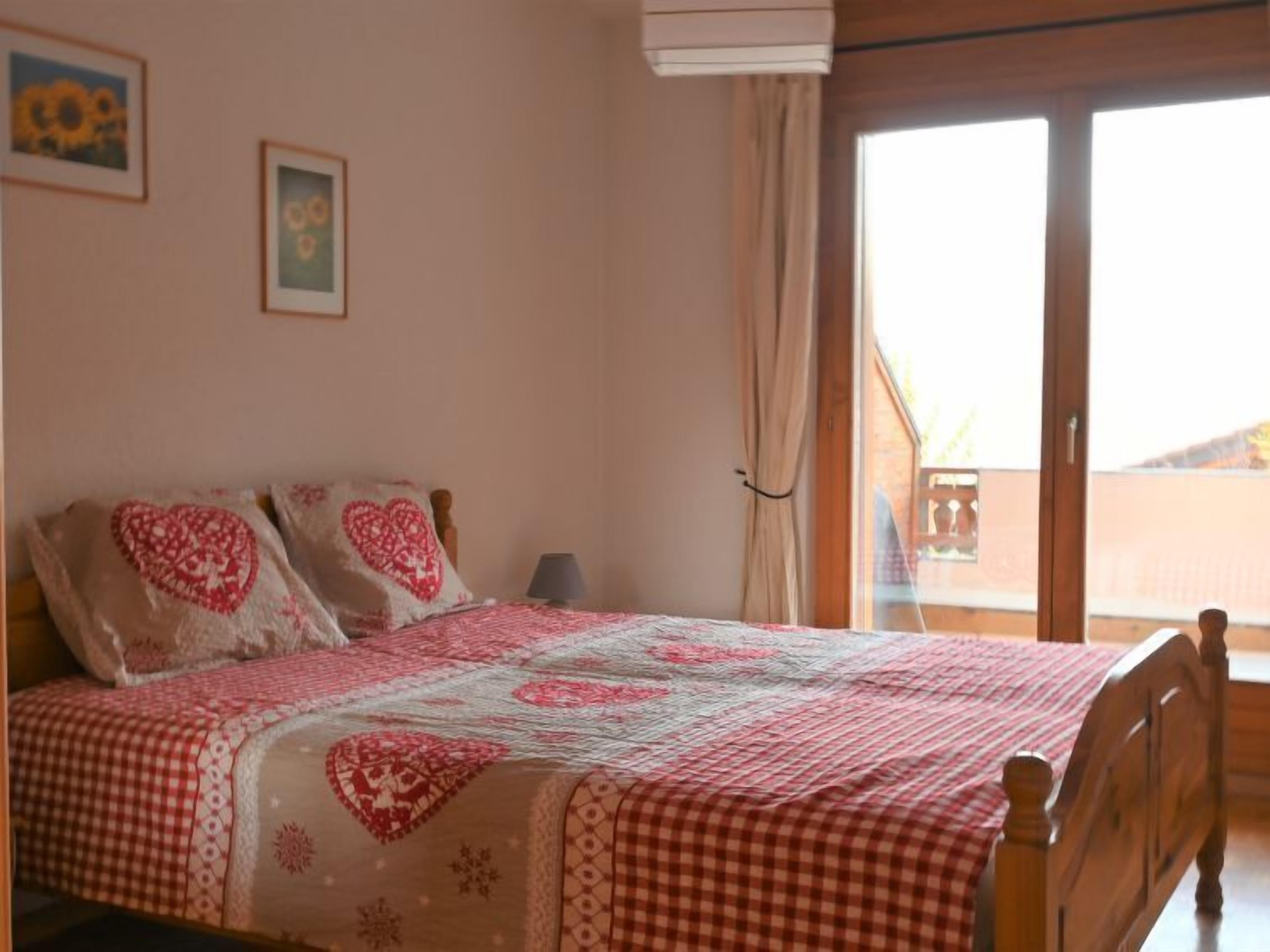 Une chambre à coucher avec un lit recouvert d’une couvre-lit à carreaux, deux coussins à motifs en forme de cœur et une fenêtre donnant sur un immeuble.