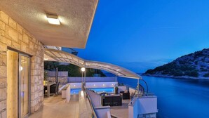Una terraza moderna al aire libre con piscina, paredes de piedra y vista al mar al anochecer.