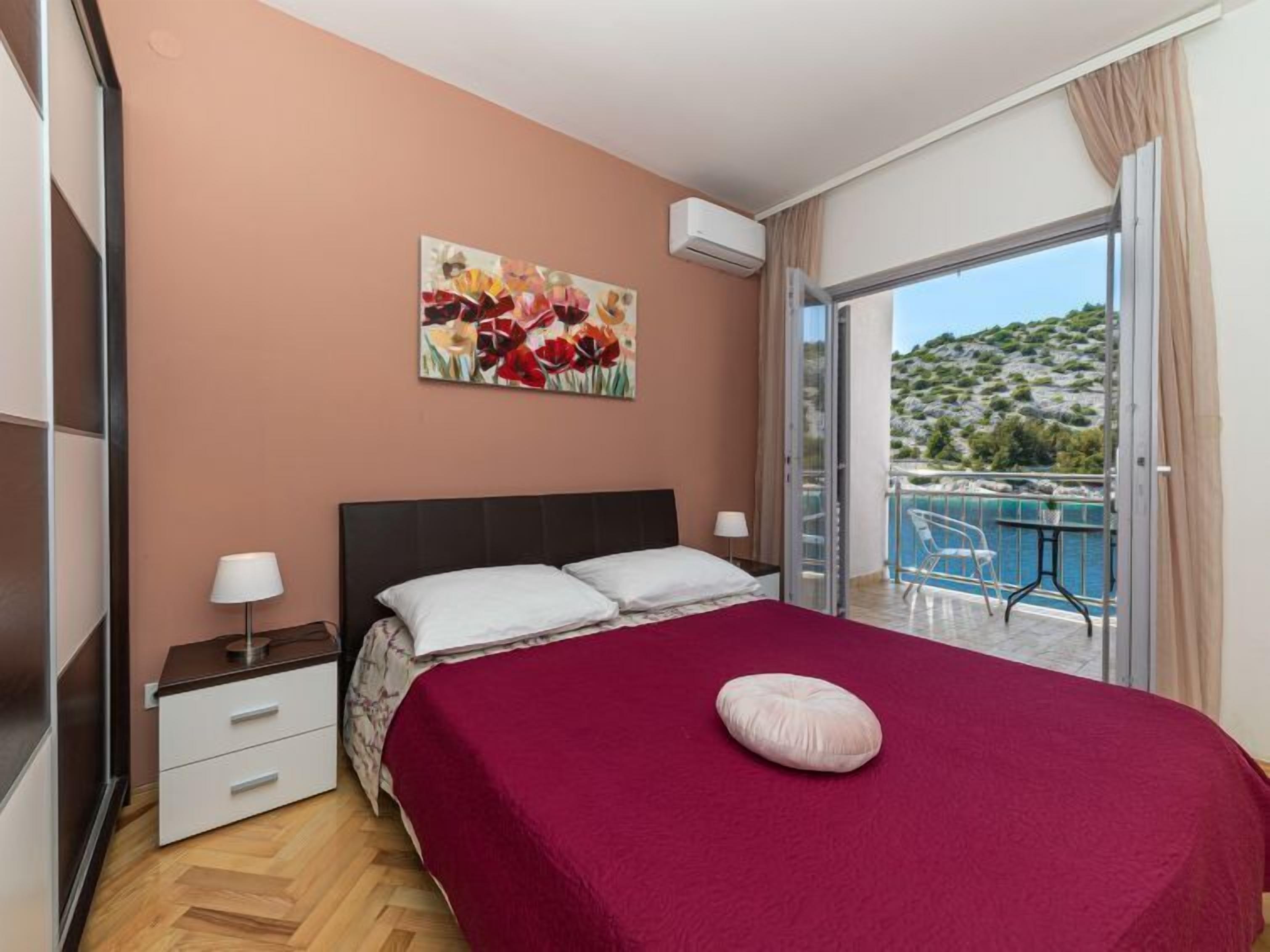 A bedroom with a bed, a nightstand, a lamp, a painting, an air conditioner, and a balcony with a view of the sea.