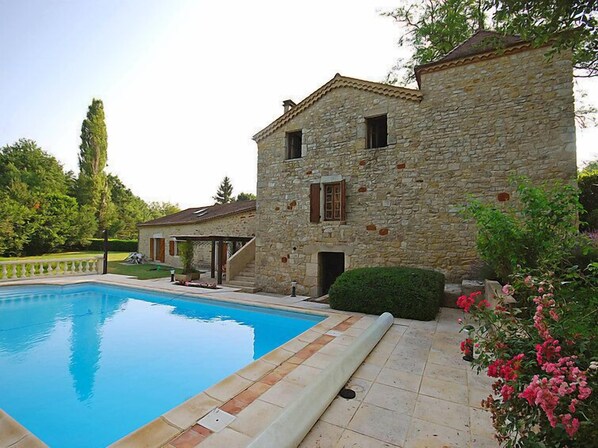 A stone building with a swimming pool, surrounded by greenery and a hedge.
