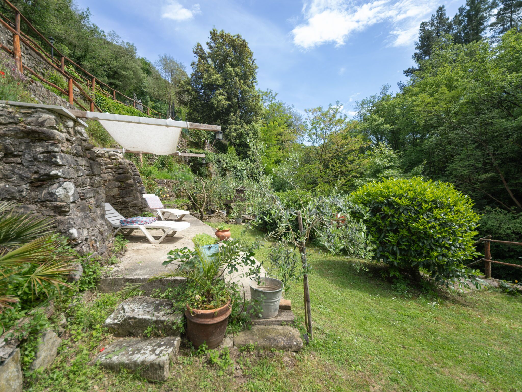 A stone wall with a wooden deck, outdoor seating, and potted plants in a lush garden setting.