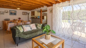 A cozy living room with a wooden ceiling, a sofa with patterned cushions, a coffee table, and a view of the outdoors through sheer curtains.