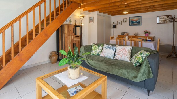 A cozy living room with a wooden staircase, a green sofa, a glass coffee table, and a dining area with a table and chairs.
