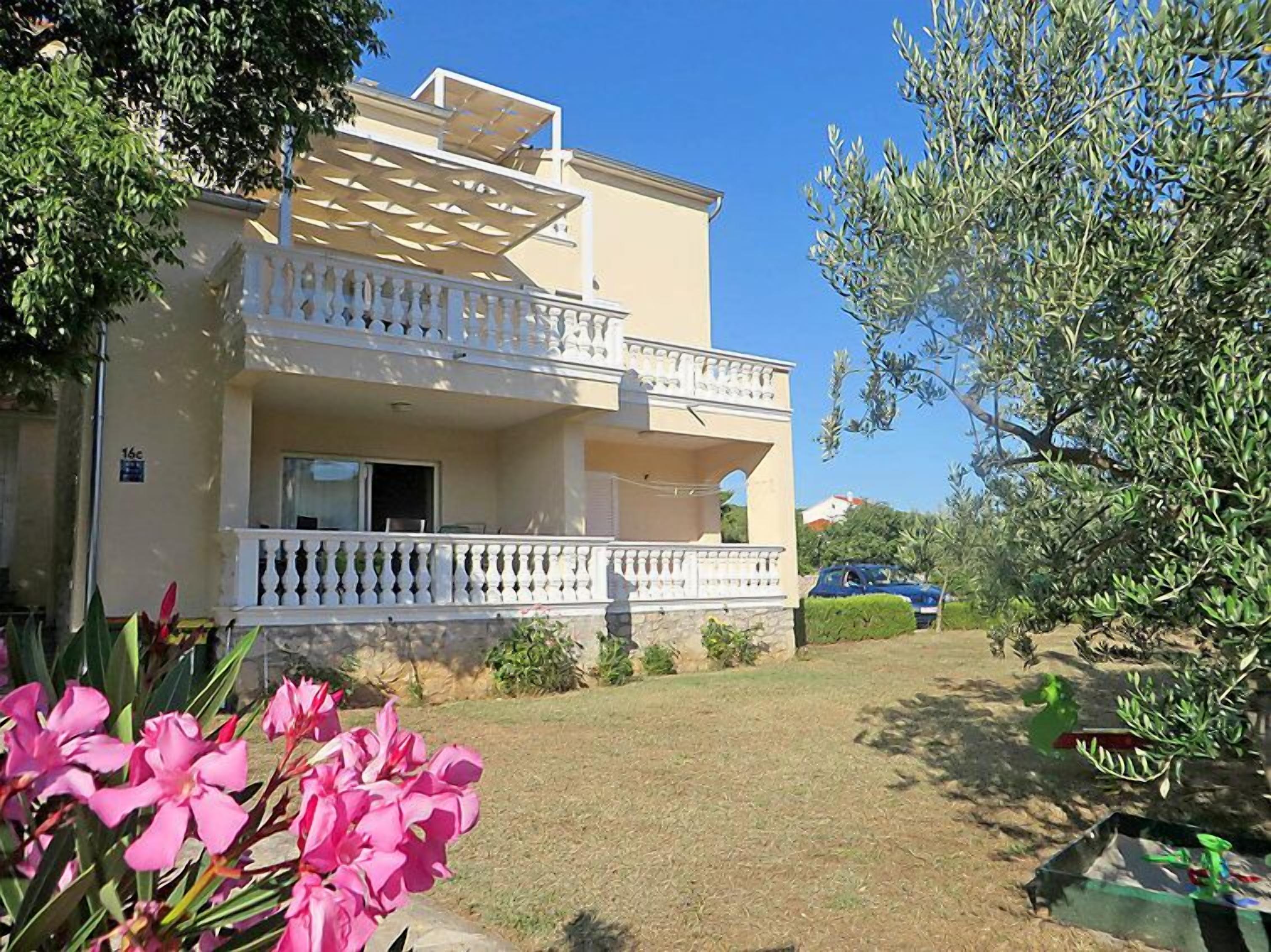 A two-story building with a balcony, a car parked in the driveway, and a garden with pink flowers.