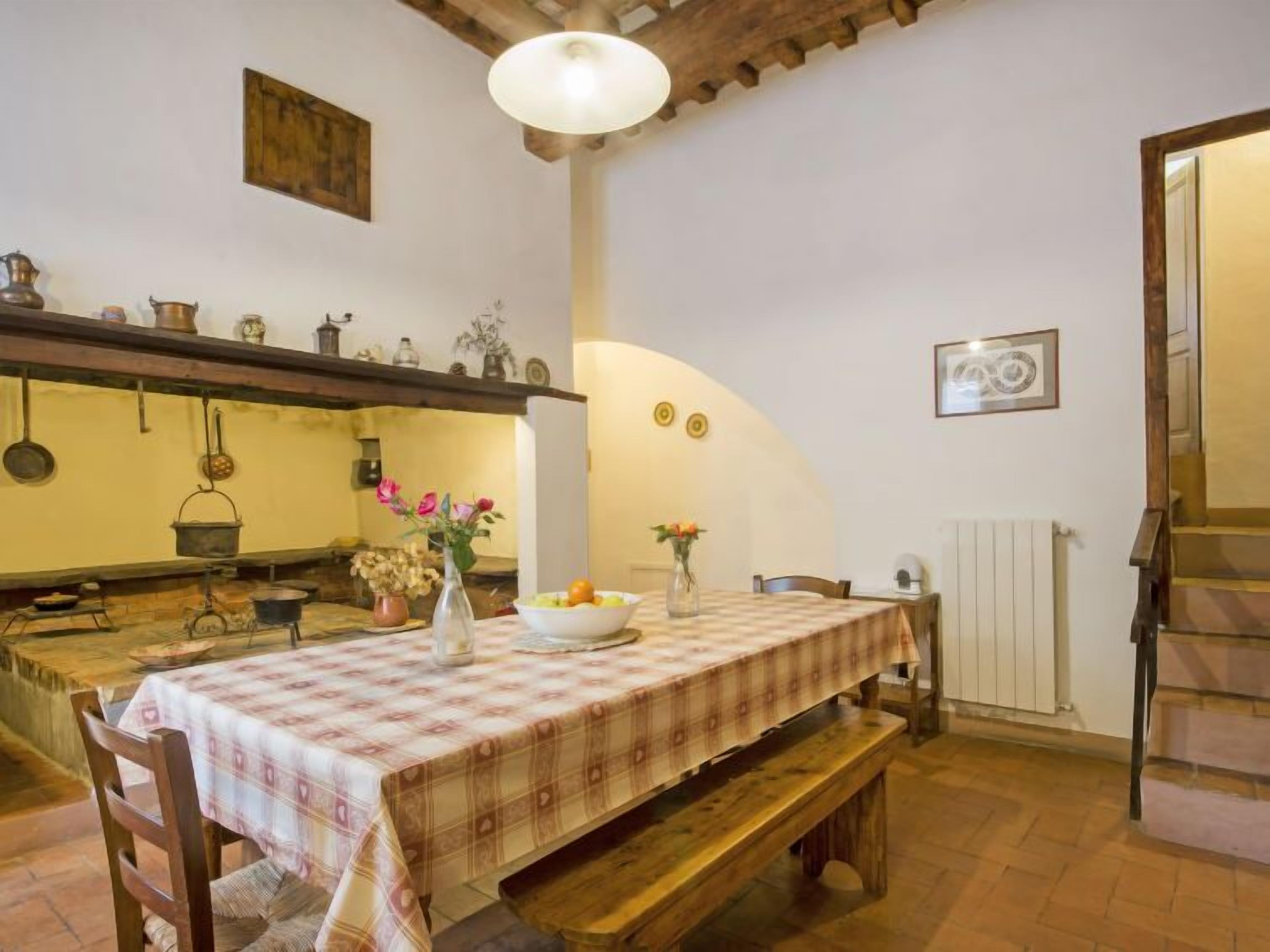 Une salle à manger rustique avec une longue table dressée pour deux personnes, des bancs en bois, un luminaire suspendu et un coin cuisine avec des casseroles et des poêles accrochées.