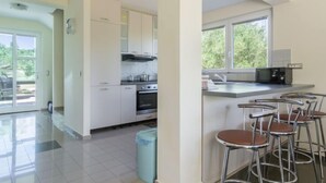 A modern kitchen with white cabinets, a built-in oven, and a microwave. There are four bar stools near a window with a view of greenery outside. - Vacation home Tea by Interhome (Pašman/Nevidane)