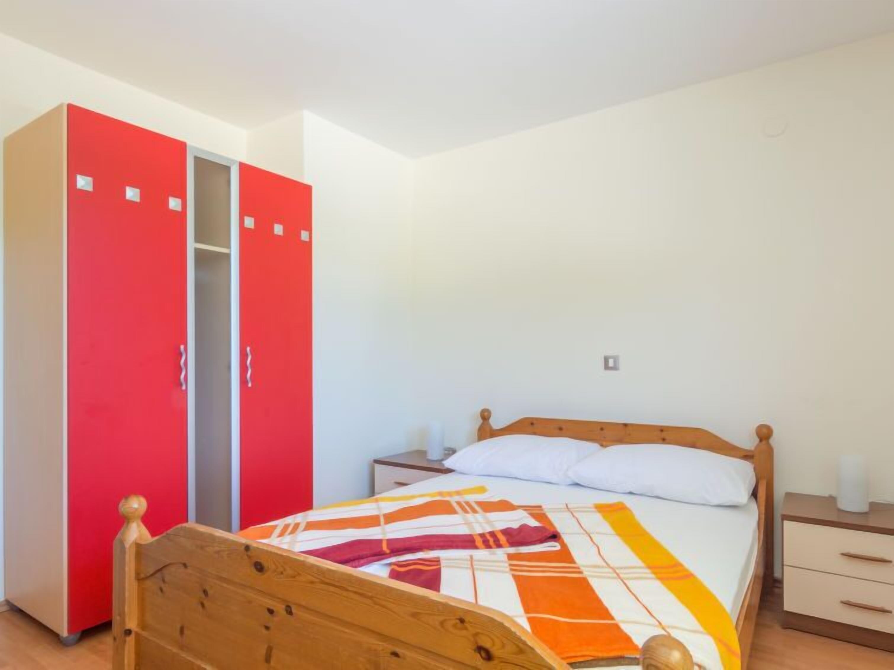 A bedroom with a wooden bed, a colorful blanket, a red wardrobe, and a bedside table with a lamp.
