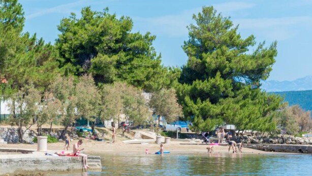A lakeside area with people relaxing by the water, surrounded by trees and a stone wall. - Vacation home Tea by Interhome (Pašman/Nevidane)