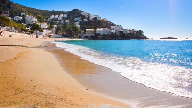 A beach with clear blue waters, a sandy shore, and buildings on a hillside in the background. - Joan Sarda by Interhome (Roses)