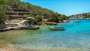 Una zona costera despejada con una playa de guijarros, un barco pequeño amarrado cerca de la orilla y una construcción de piedra con asientos al aire libre.