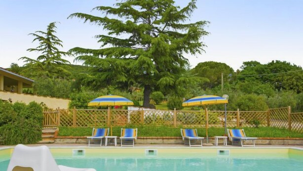 Una piscina con sillas de descanso y sombrillas, rodeada por un muro de piedra y una valla de madera.