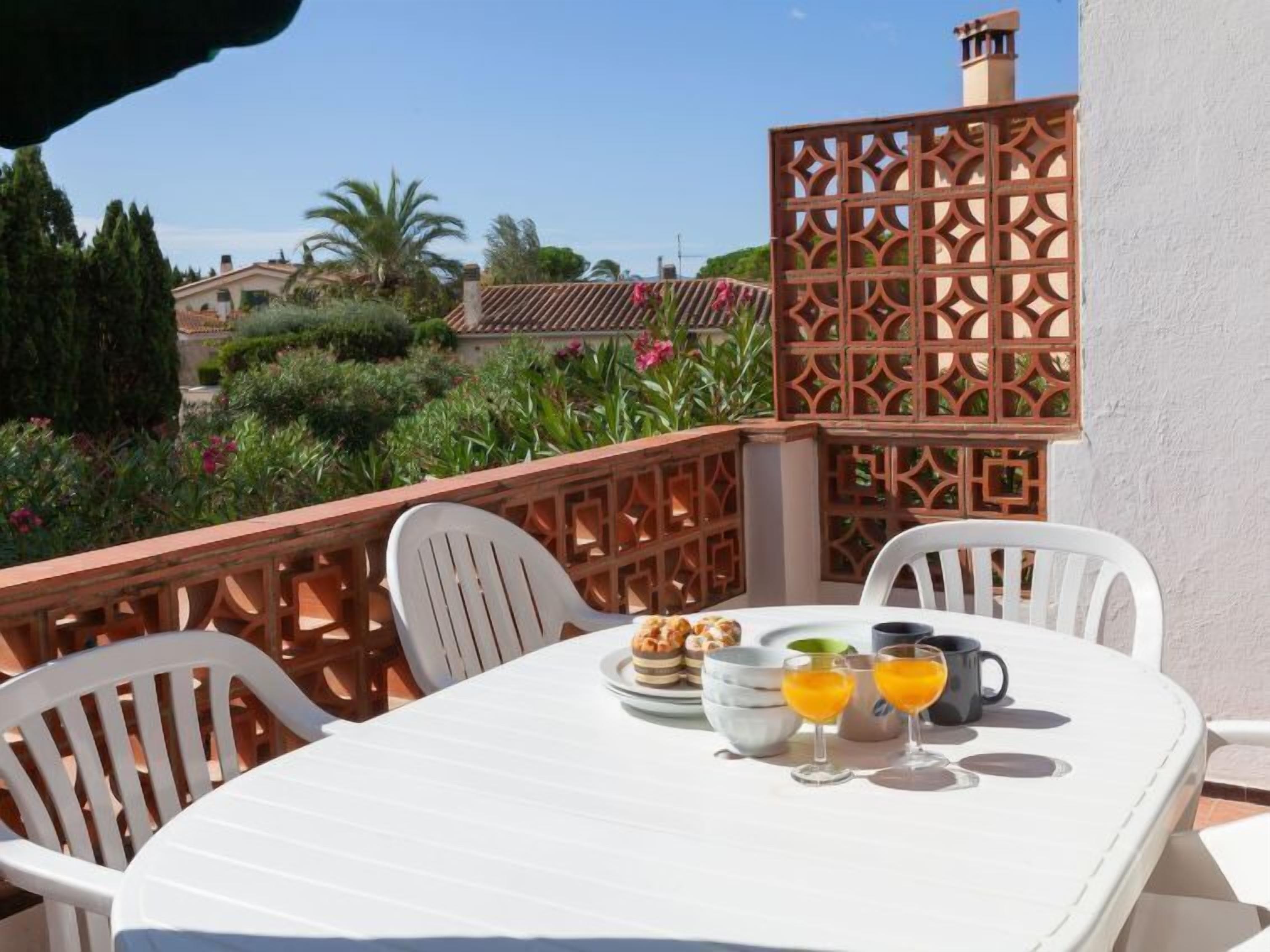Una mesa blanca con sillas en un balcón, una valla de madera decorativa y vistas a casas y follaje.