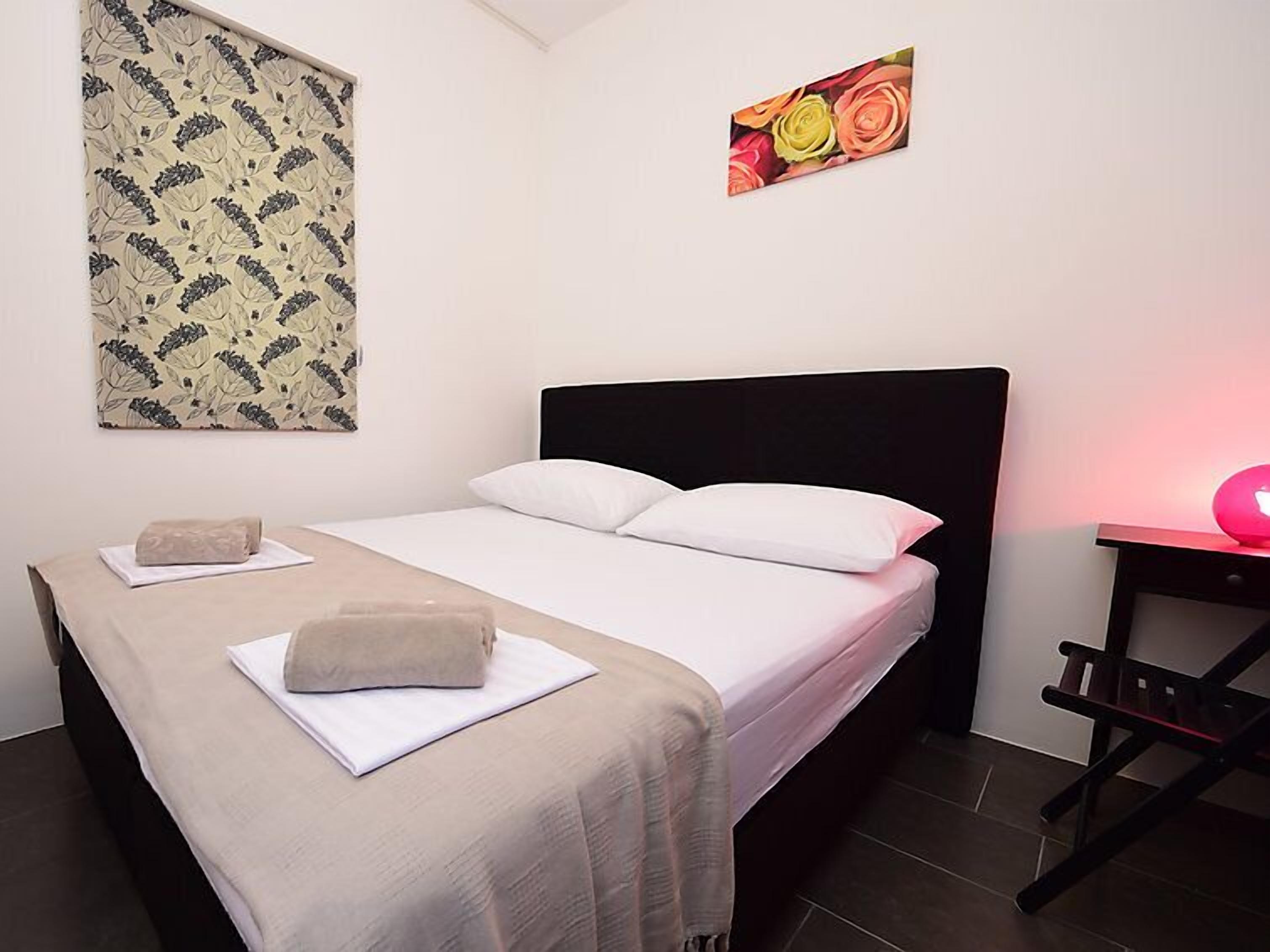 A neatly made bed with a white and beige bedspread, a black headboard, and a single pillow. A small bedside table with a pink lamp is visible to the right.