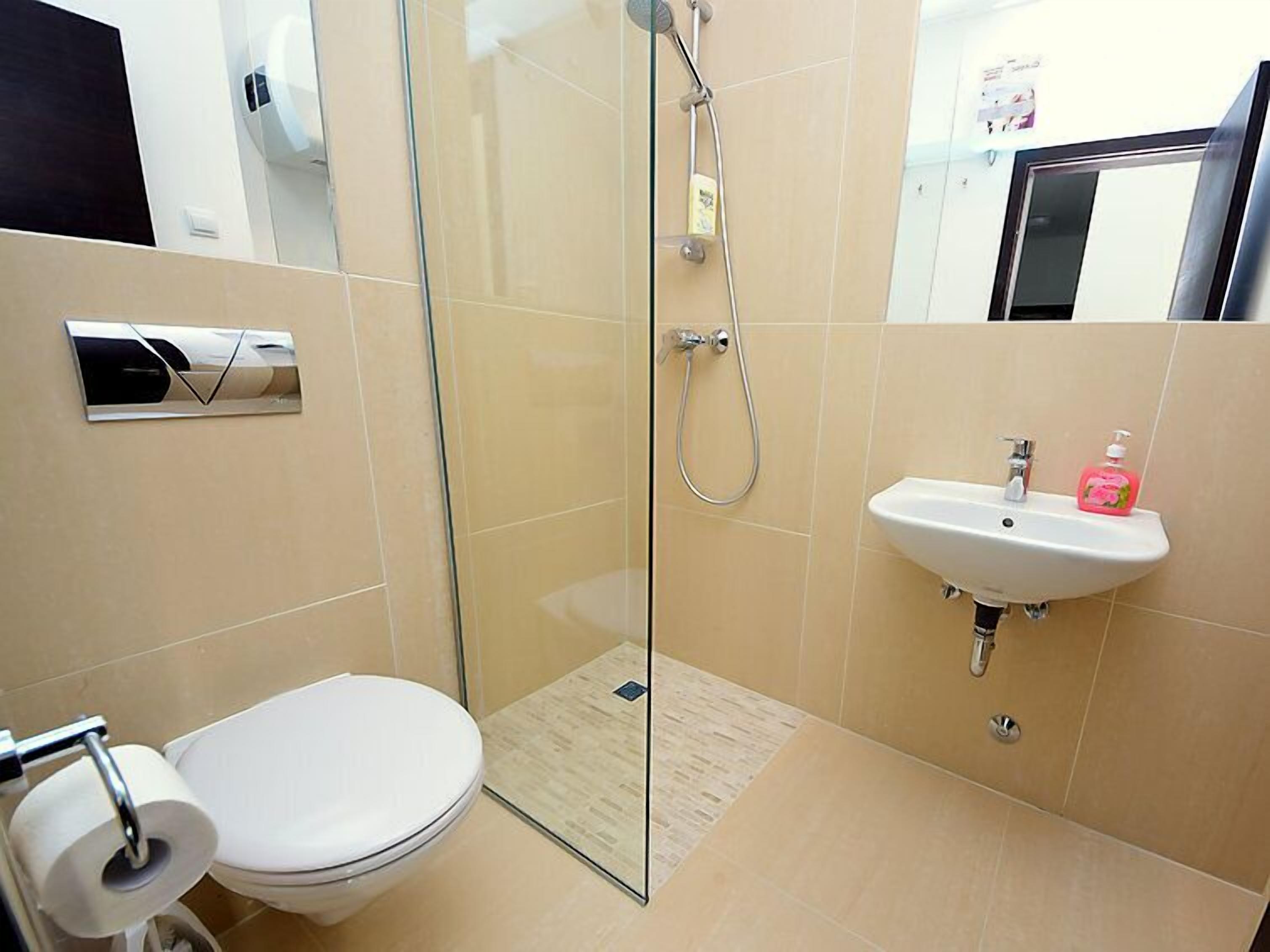 Une salle de bain moderne avec une douche à parois de verre, des toilettes blanches, un lavabo avec un distributeur de savon et des murs recouverts de carreaux beiges.