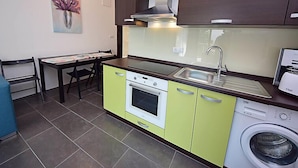 Een kleine keuken met een fornuis, spoelbak en vaatwasser, een eettafel met stoelen en een schilderij aan de muur.