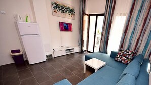 Een moderne woonkamer met een blauwe bank, een klein wit tafeltje, en een keukenhoek met een witte koelkast en een afvalbak.
