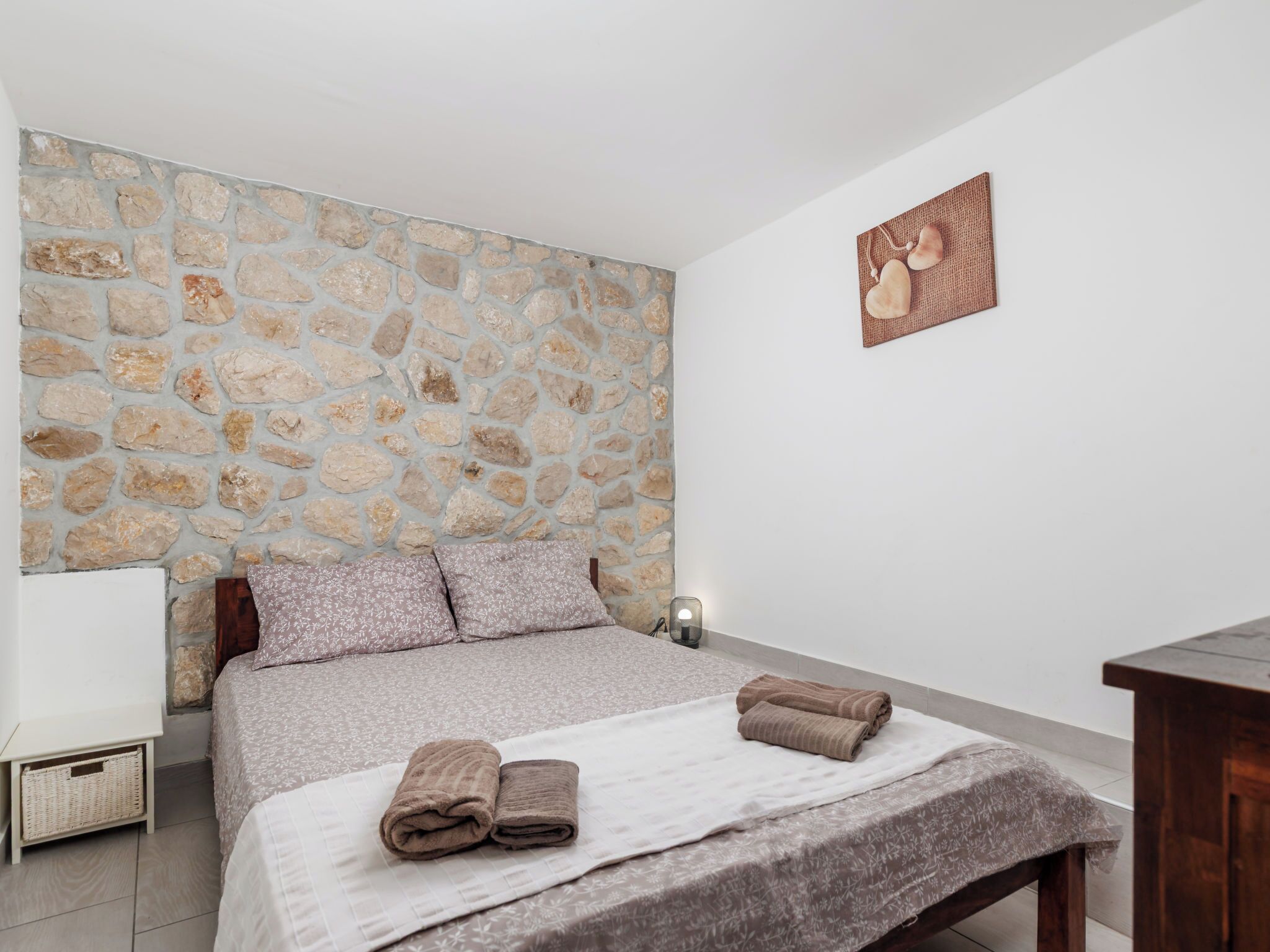 A bedroom with a stone wall, a bed with a white and brown patterned bedspread, two pillows, and two rolled towels on the bed.