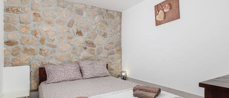 A bedroom with a stone wall, a bed with a white and brown patterned bedspread, two pillows, and two rolled towels on the bed.