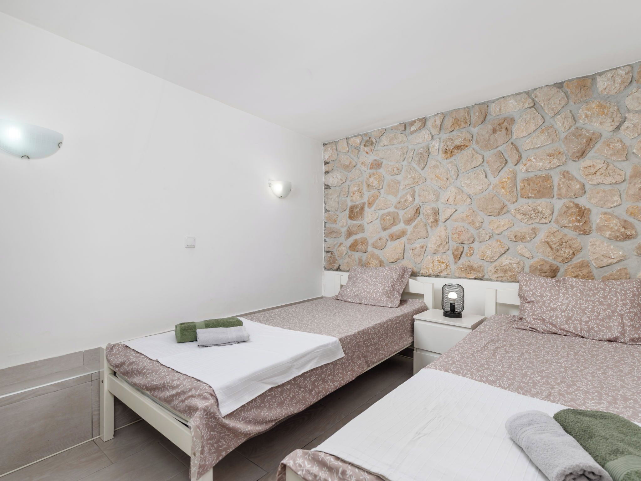 Un dormitorio con dos camas individuales, una pared de piedra, una mesita de noche con lámpara y una lámpara de techo blanca.