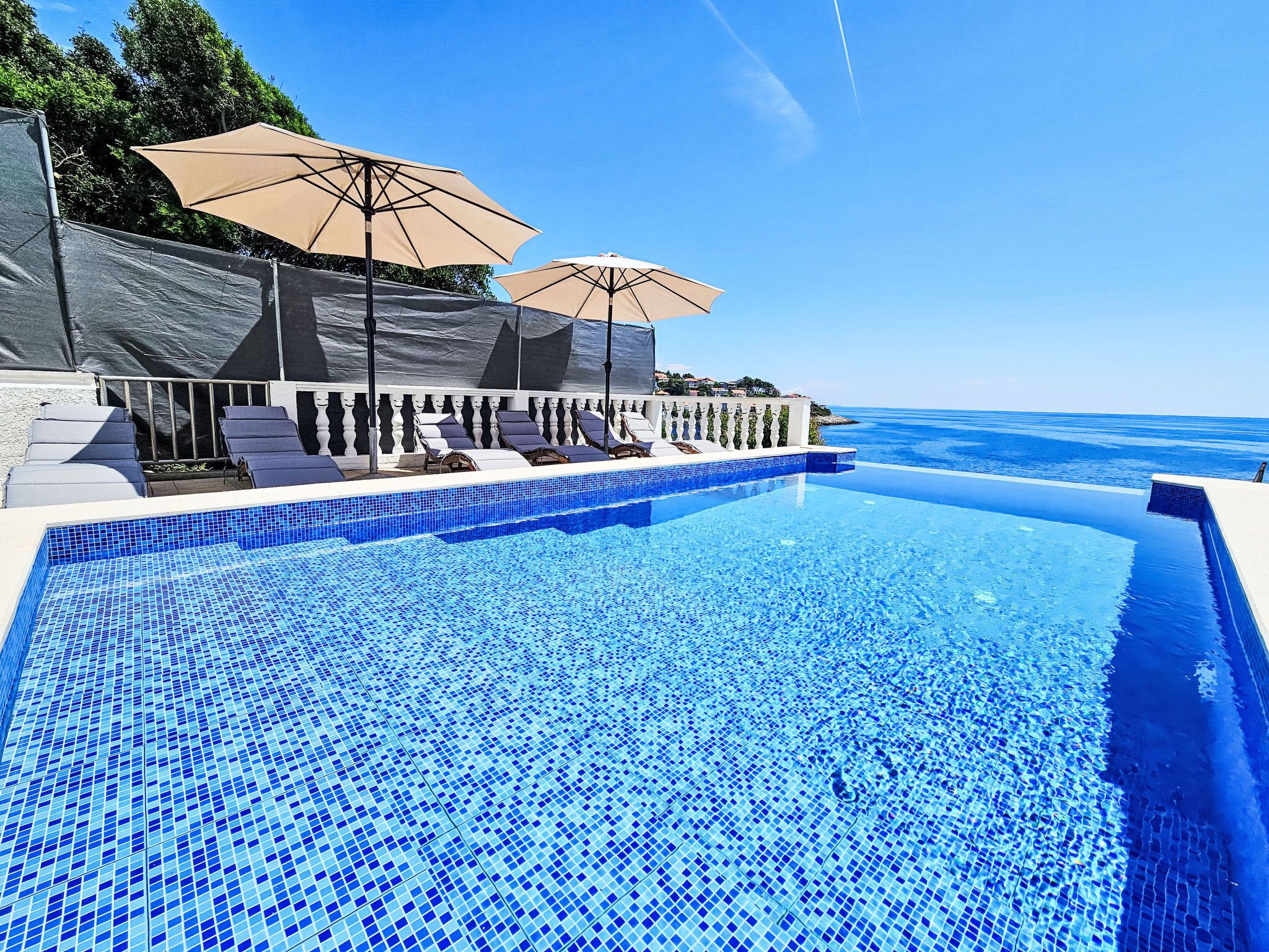 A clear blue infinity pool with lounge chairs and umbrellas, overlooking a calm sea.