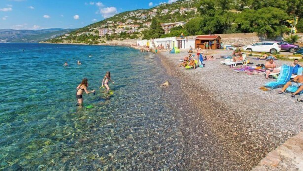 Una playa con aguas poco profundas y transparentes, gente nadando y tomando el sol, y casas al fondo.