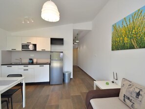 A modern kitchen with white cabinets, a microwave, and a stainless steel refrigerator. There is a dining table with chairs, a small white table with a green object, and a brown sofa with a patterned cushion. A painting of yellow flowers hangs on the wall. - Apartment Ivka by Interhome (Vodice/Tribunj)