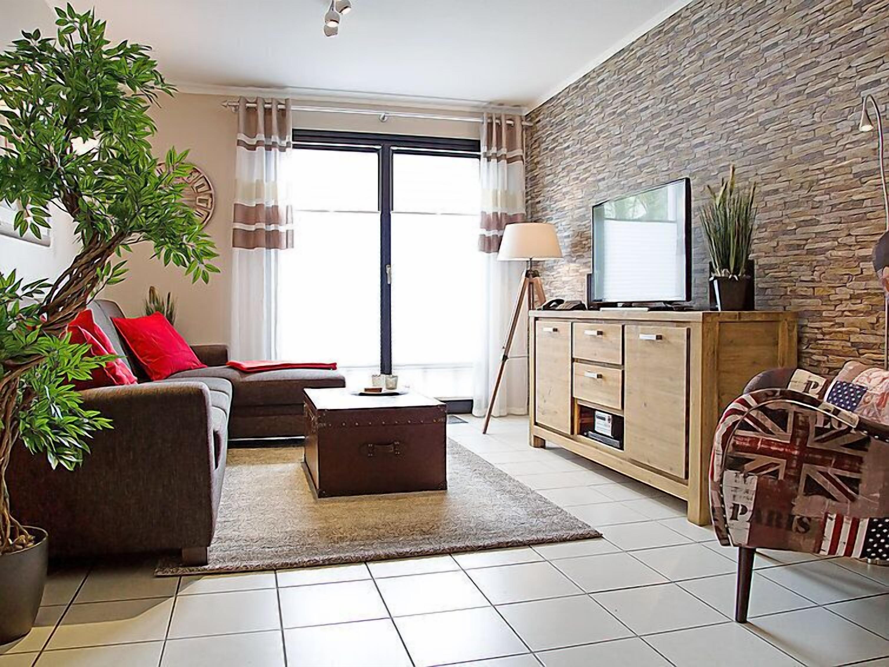 A modern living room with a sofa, coffee table, and a television on a wooden cabinet. There is a large window with curtains, a potted plant, and a lamp.