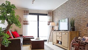 Un saló modern amb un sofà, una taula de centre i una televisió sobre un moble de fusta. Hi ha una finestra gran amb cortines, una planta en test i una làmpada.