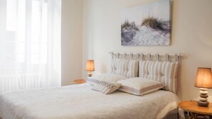 Quarto com cama, duas luminárias de cabeceira, janela com cortinas e um quadro na parede.