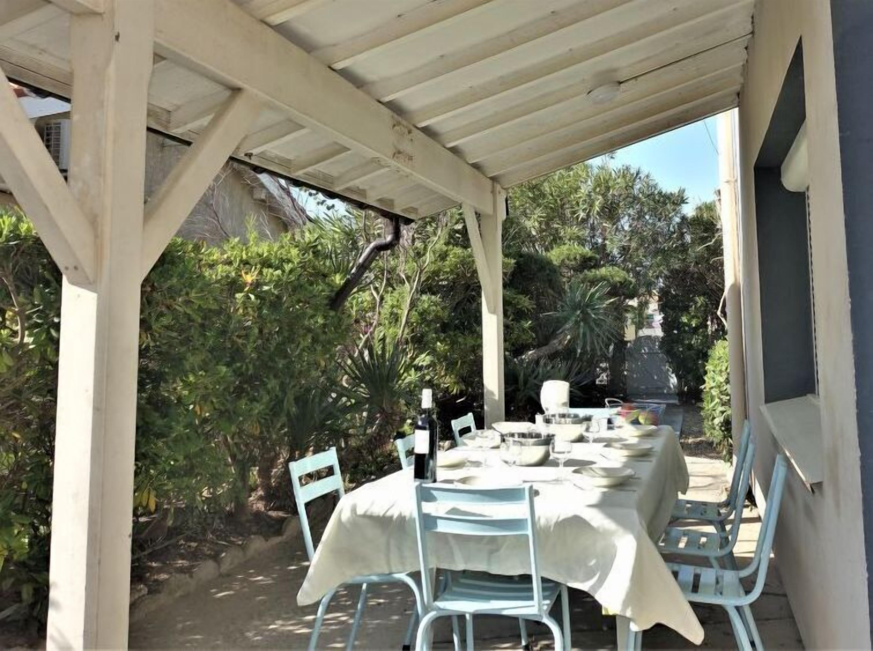 Une terrasse extérieure couverte, avec une table dressée pour le repas, entourée de verdure et sous un ciel dégagé.