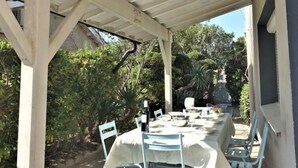 Eine überdachte Terrasse im Freien mit Esstisch, umgeben von Pflanzen und blauem Himmel.