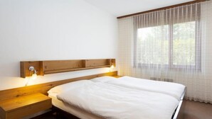 A neatly arranged bedroom with a wooden headboard, a single bed, bedside tables with lamps, and a large window with sheer curtains. - Resort al Centro by Interhome (Breganzona)